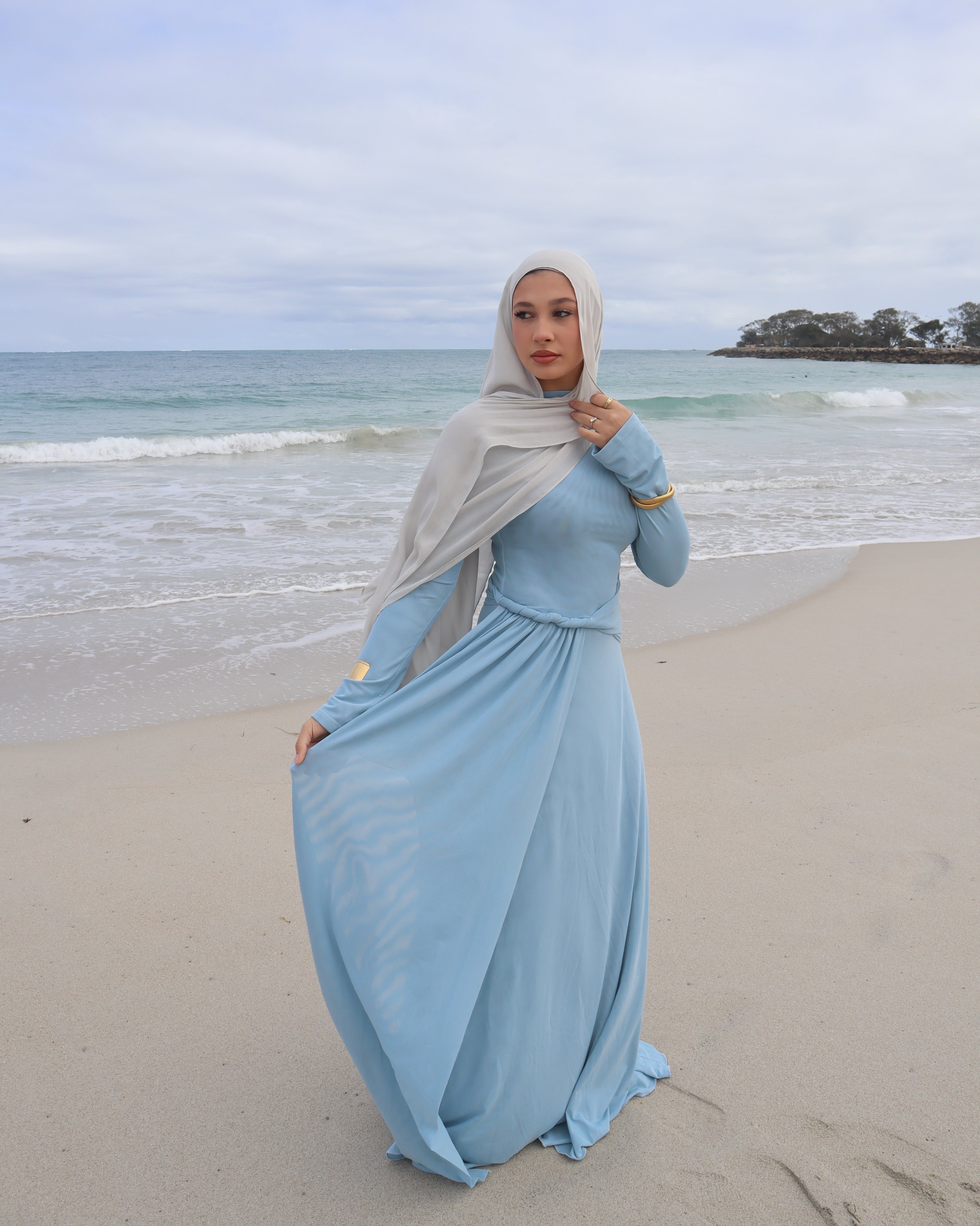 Woman in a long light blue dress standing on a sandy beach with ocean in the background