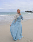 Woman in a long light blue dress standing on a sandy beach with ocean in the background